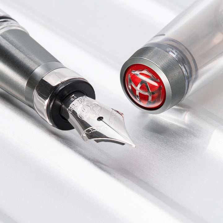 Detailed close-up of the TWSBI Diamond 580ALR Nickel Gray fountain pen lying on a smooth metallic surface. The shot tightly frames the silver stainless steel Jowo nib and ribbed matte nickel-gray grip section, next to the detached clear cap showcasing the signature red TWSBI finial logo.