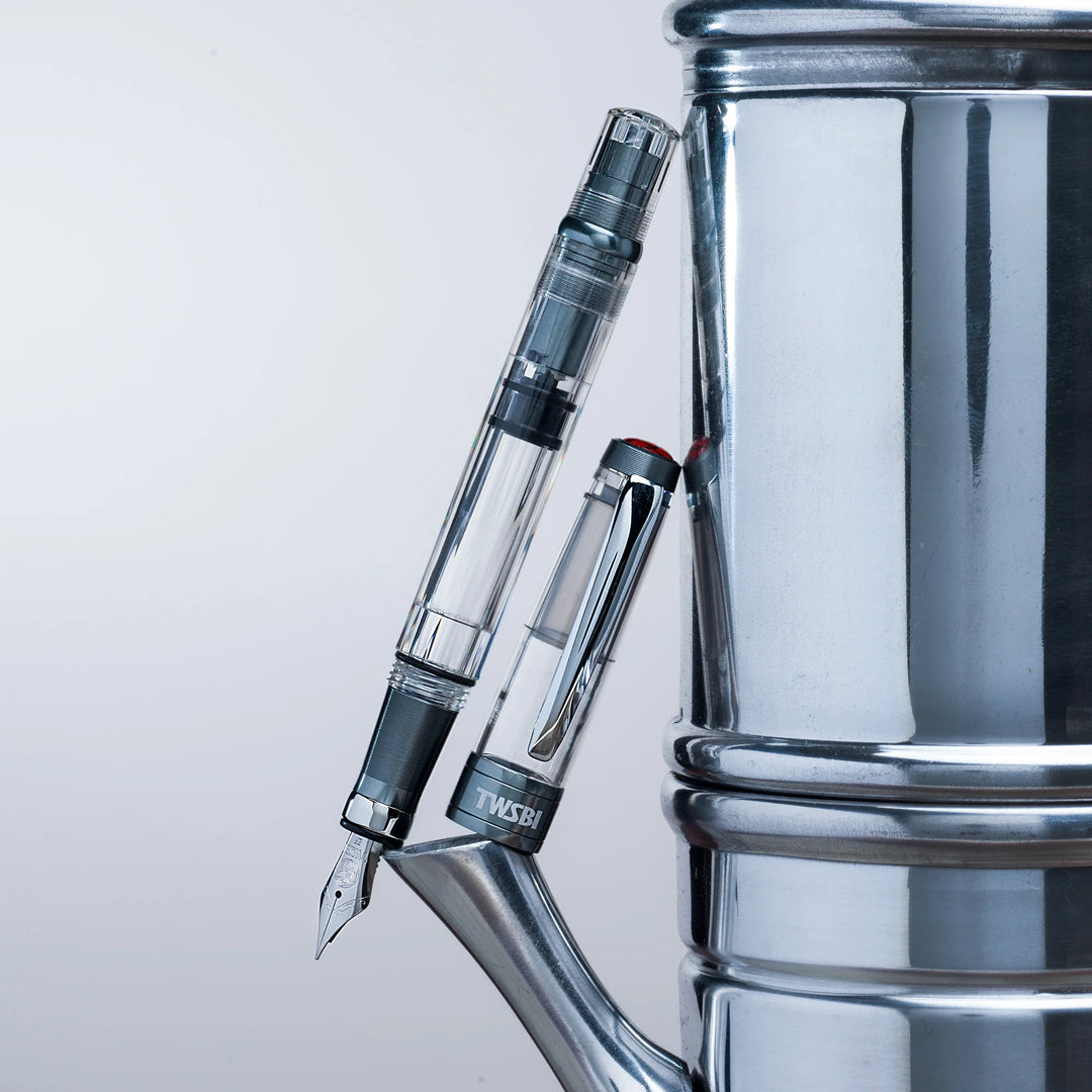 Creative, industrial product shot showing the uncapped TWSBI Diamond 580ALR Nickel Gray fountain pen and cap leaning vertically against a polished stainless steel espresso maker or kettle. The cool metallic environment perfectly complements the pen's matte nickel-gray aluminum trim and clear demonstrator design.