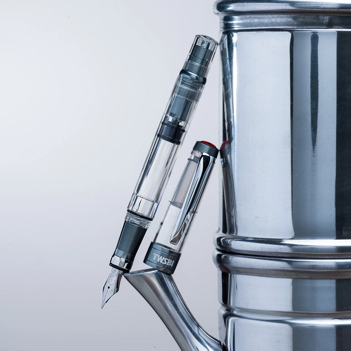 Creative, industrial product shot showing the uncapped TWSBI Diamond 580ALR Nickel Gray fountain pen and cap leaning vertically against a polished stainless steel espresso maker or kettle. The cool metallic environment perfectly complements the pen's matte nickel-gray aluminum trim and clear demonstrator design.