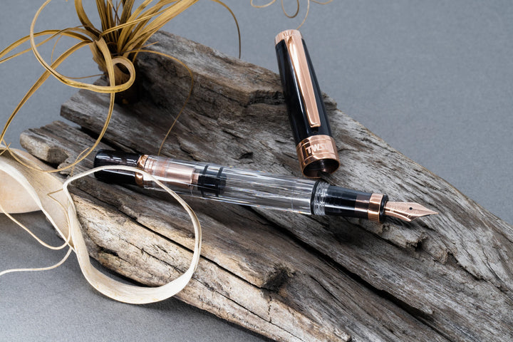 Uncapped TWSBI Diamond 580 Smoke RoseGold II fountain pen styled on weathered wood, displaying the clear ink barrel, internal rose gold piston filling mechanism, and matching smoke resin cap with a rose gold clip.
