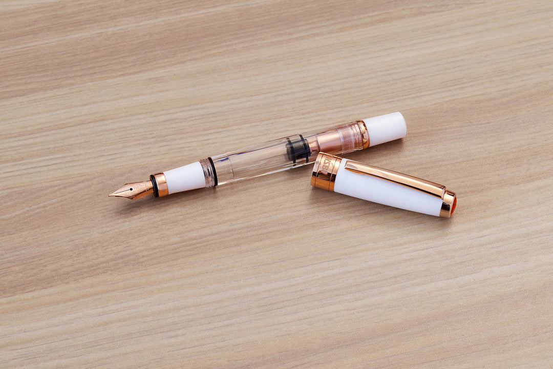 Uncapped TWSBI Diamond 580 White Rose Gold II fountain pen styled on light wood grain, showcasing the clear demonstrator barrel, rose gold piston mechanism, and matching solid white resin cap.