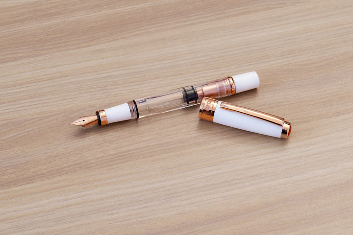 Uncapped TWSBI Diamond 580 White Rose Gold II fountain pen styled on light wood grain, showcasing the clear demonstrator barrel, rose gold piston mechanism, and matching solid white resin cap.
