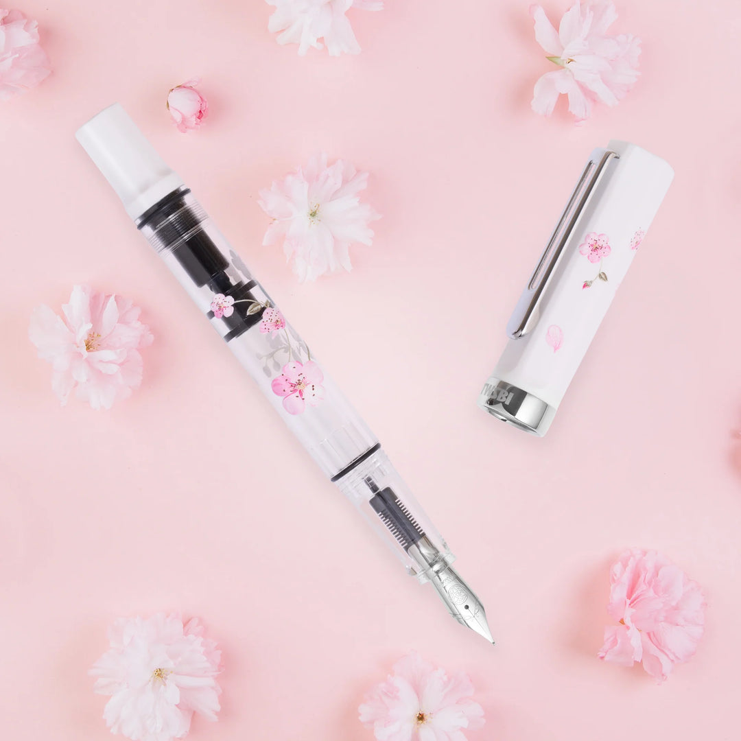 Uncapped TWSBI ECO Cherry Blossom fountain pen and matching white cap, laid flat on a pastel pink background surrounded by real pink cherry blossoms.