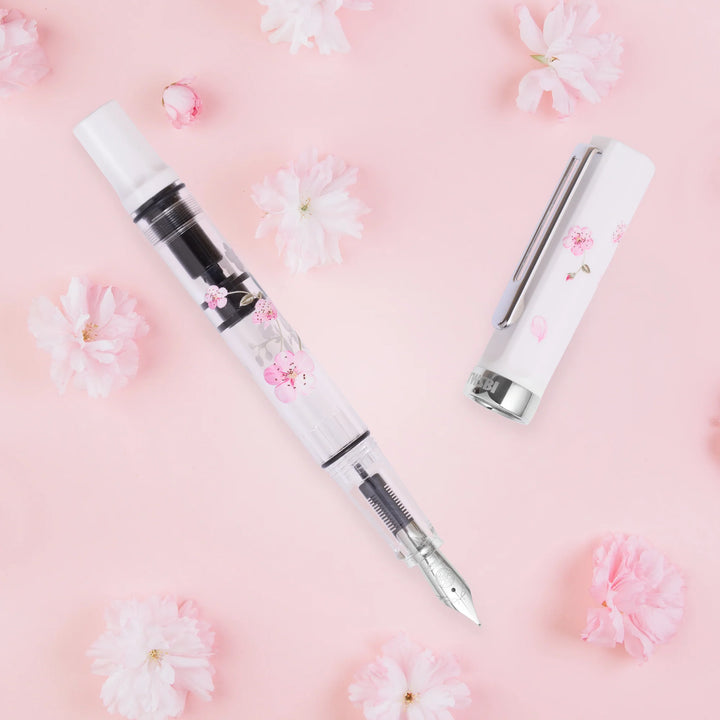 Uncapped TWSBI ECO Cherry Blossom fountain pen and matching white cap, laid flat on a pastel pink background surrounded by real pink cherry blossoms.