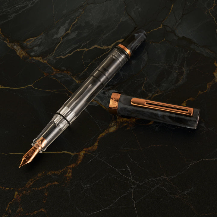 Top-down view of the uncapped TWSBI ECO Marble Bronze fountain pen and its cap resting on a dark, gold-veined marble surface. The image highlights the vintage bronze-plated nib, the crystal-clear demonstrator barrel, and the swirled black marble resin cap with its matching bronze pocket clip.