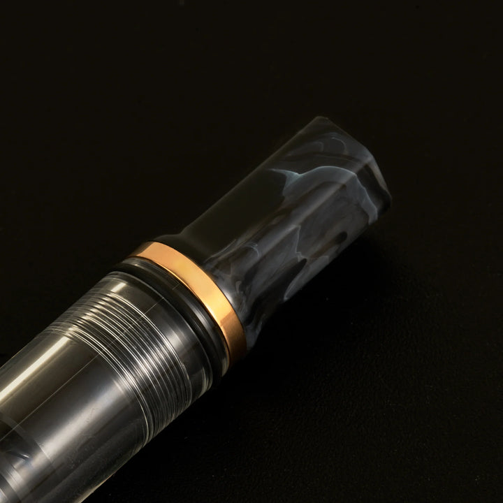 Macro close-up of the TWSBI ECO Marble Bronze fountain pen against a solid black background, focusing tightly on the piston knob. The image details the distinct swirled black and gray marble resin, the polished bronze-plated accent ring, and the clear barrel revealing the internal piston mechanism.