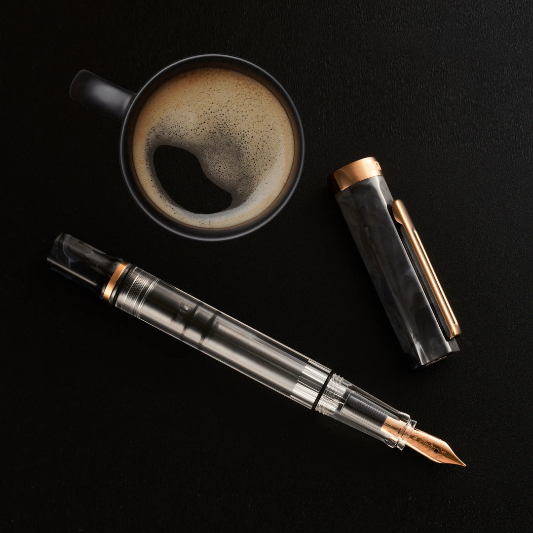 Cozy, top-down flat lay photograph featuring the uncapped TWSBI ECO Marble Bronze fountain pen and its cap arranged next to a black ceramic mug filled with fresh, frothy espresso. The dark background perfectly complements the pen's black marble resin and warm bronze-plated accents.