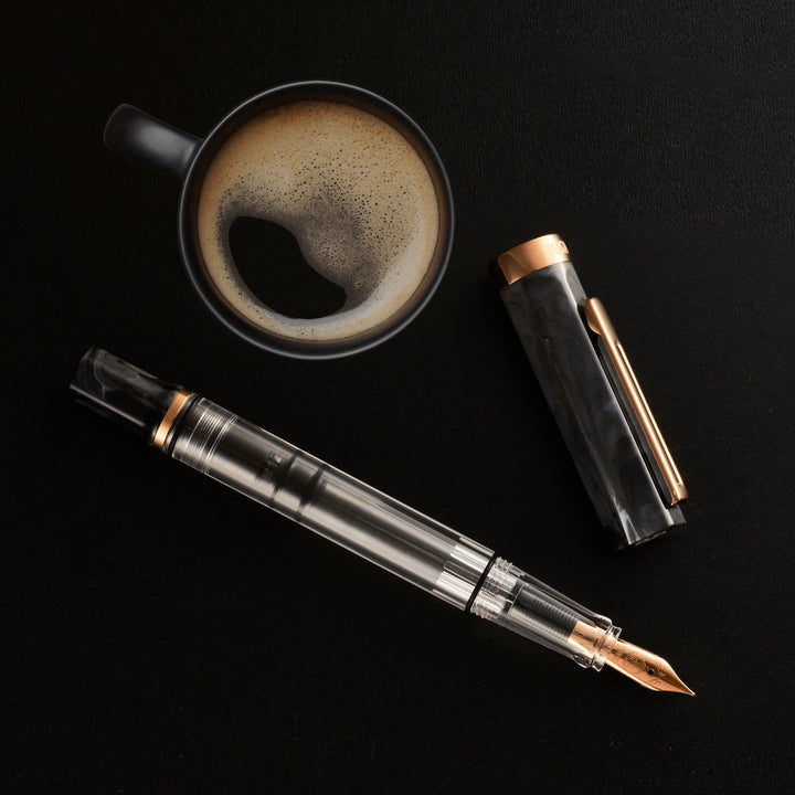 Cozy, top-down flat lay photograph featuring the uncapped TWSBI ECO Marble Bronze fountain pen and its cap arranged next to a black ceramic mug filled with fresh, frothy espresso. The dark background perfectly complements the pen's black marble resin and warm bronze-plated accents.
