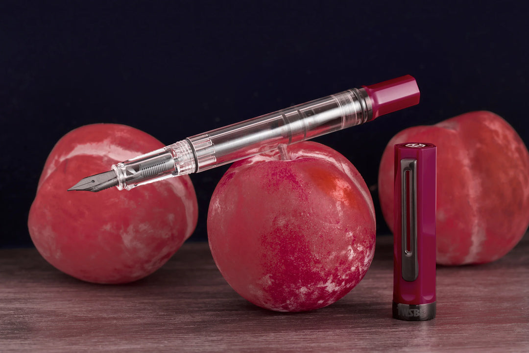 Moody product photograph of the uncapped TWSBI ECO Plum Onyx fountain pen resting horizontally across a fresh red plum, with its matching deep purple cap standing vertically beside it. The dark background and rustic wooden surface highlight the pen's clear demonstrator barrel and stealthy dark onyx-plated nib and trim.