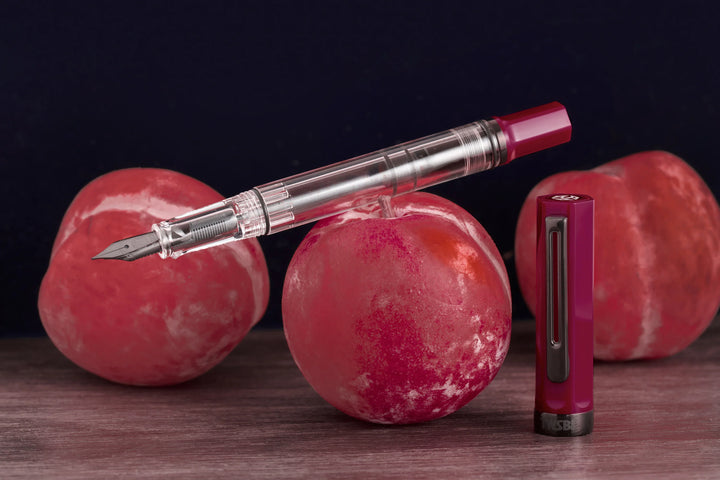 Moody product photograph of the uncapped TWSBI ECO Plum Onyx fountain pen resting horizontally across a fresh red plum, with its matching deep purple cap standing vertically beside it. The dark background and rustic wooden surface highlight the pen's clear demonstrator barrel and stealthy dark onyx-plated nib and trim.