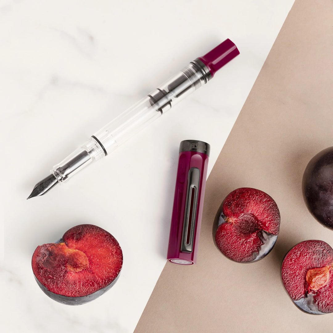 Top-down flat lay photograph of the uncapped TWSBI ECO Plum Onyx fountain pen and its cap resting on white marble, beautifully contrasted alongside sliced and whole fresh dark plums on a warm beige surface. The elegant composition emphasizes the rich plum purple resin and sleek onyx black accents.
