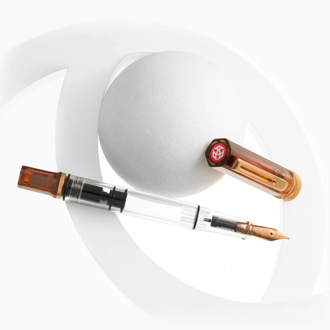 Top-down view of the uncapped TWSBI ECO Rootbeer Bronze fountain pen and its detached cap arranged diagonally over a white textured sphere and subtle display rings on a pure white background. The shot details the bronze-plated nib, the internal piston mechanism, and the signature red TWSBI finial logo on the cap.