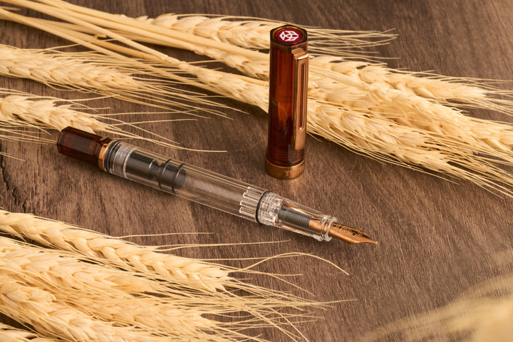 Warm lifestyle photograph of the uncapped TWSBI ECO Rootbeer Bronze fountain pen and its vertically standing cap resting on a dark wood grain surface next to stalks of dried golden wheat. The natural setting beautifully highlights the vintage bronze-plated nib, the clear demonstrator barrel, and the rich translucent brown rootbeer resin.