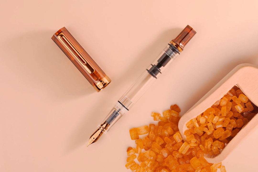 Top-down flat lay photograph of the uncapped TWSBI ECO Rootbeer Bronze fountain pen and its cap on a soft peach background, styled next to a wooden scoop overflowing with amber rock sugar crystals. The composition emphasizes the warm translucent brown resin and the matching vintage bronze-plated pocket clip and trim.