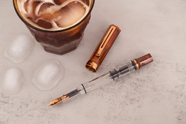 Refreshing lifestyle photograph of the uncapped TWSBI ECO Rootbeer Bronze fountain pen and its cap laid on a textured light grey stone surface alongside melting ice cubes and a chilled glass of iced coffee. The shot showcases the rich brown rootbeer resin piston knob and cap paired perfectly with vintage bronze accents.