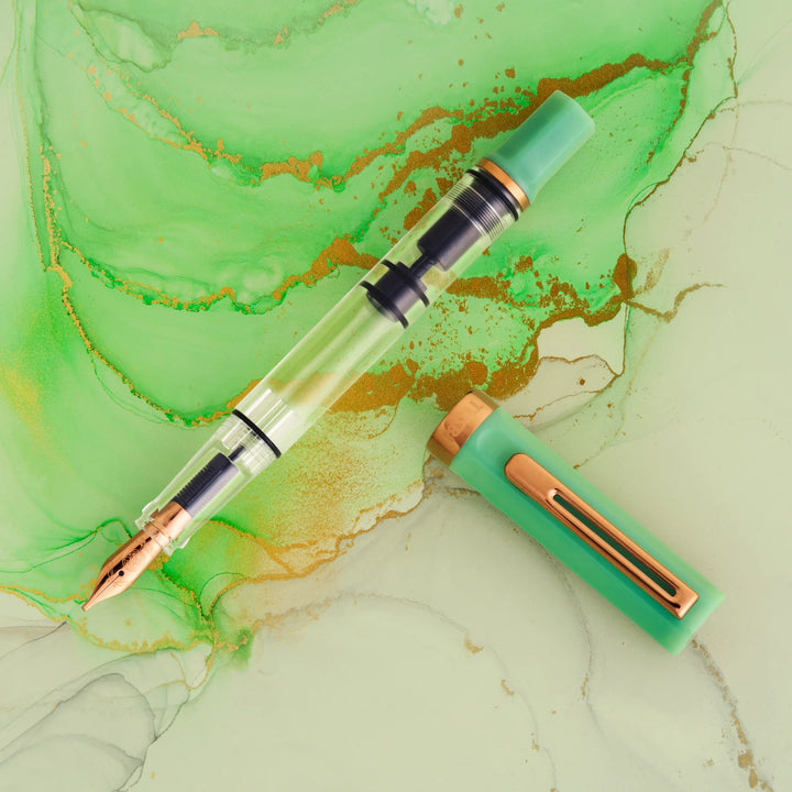 Uncapped TWSBI ECO Jade Jelly Bronze fountain pen showing the clear demonstrator barrel and bronze-coloured nib, laid on a green and gold ink-wash background.
