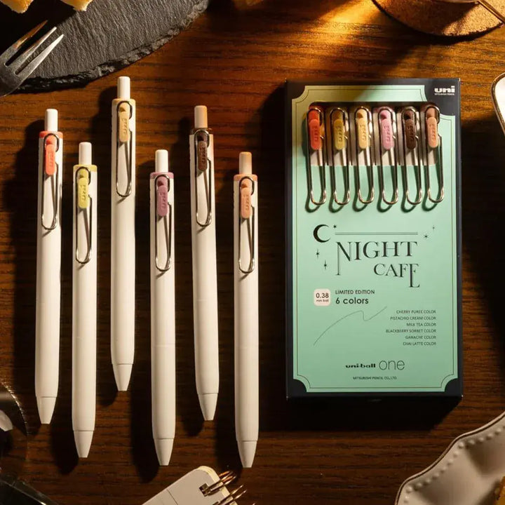 Lifestyle flat lay of Uni-ball One Night Cafe gel pens on a dark table.