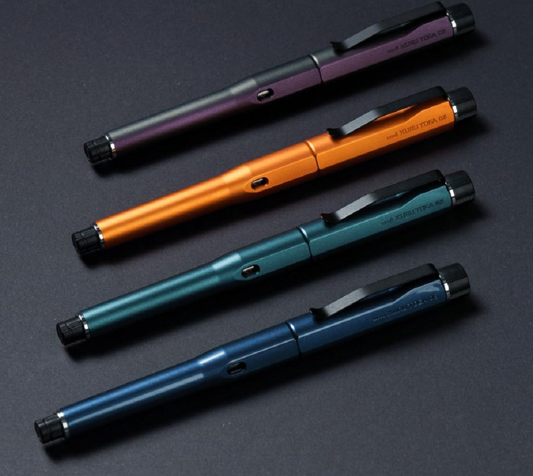 Angled view of the four Uni Kuru Toga DIVE mechanical pencil colour variants—Aurora Purple, Twilight Orange, Dense Green, and Abyss Blue—resting on a dark surface to highlight their coated ABS resin finish.
