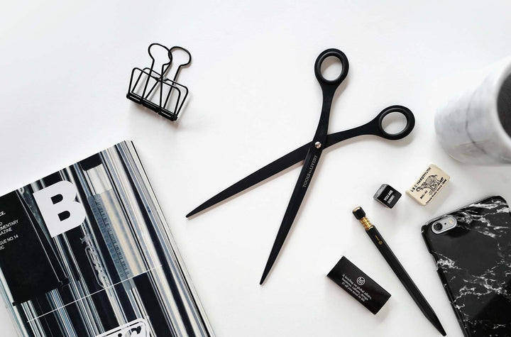 Overhead flat lay showcasing the TOOLS to LIVEBY 9-inch matte black scissors resting open on a white workspace, surrounded by premium monochrome everyday carry gear including a mechanical pencil, marble phone case, and design magazine.
