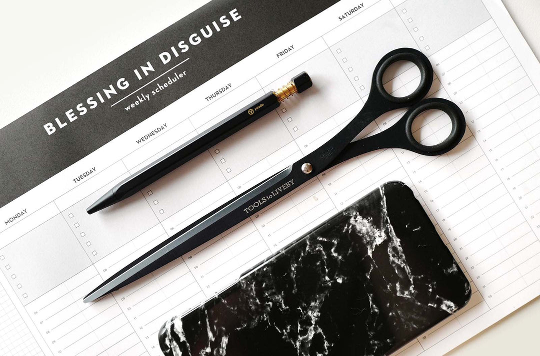Premium workspace styling featuring the TOOLS to LIVEBY 9-inch matte black scissors resting diagonally on a minimalist weekly scheduler, placed beside a brass mechanical pencil and a black marble phone case.