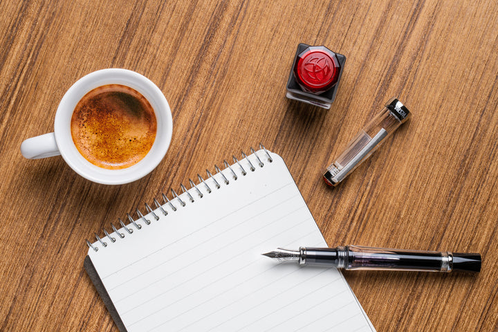 Cup of coffee, inkwell, TWSBI Vac700R Fountain Pen - Clear with filled ink, and notebook on a wooden surface