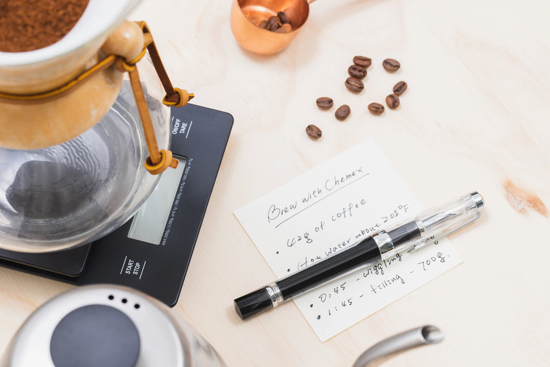 Coffee brewing equipment with coffee beans and TWSBI Vac700R Fountain Pen - Clear with filled ink on a white surface