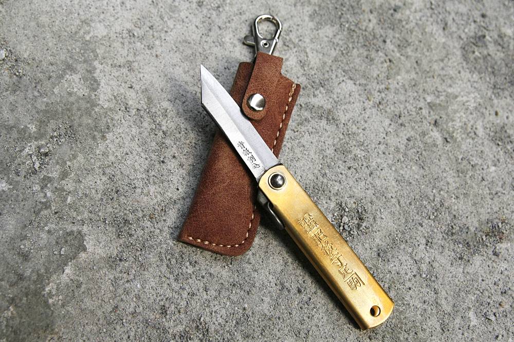 Higonokami Brass S Japanese folding knife resting open on its carry pouch against a rustic concrete background.